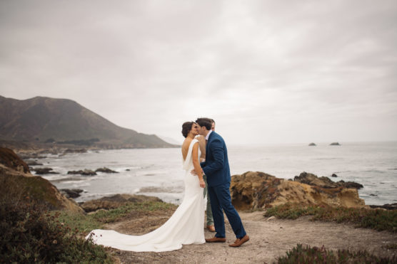 A bride and groom share a kiss near a rocky coastline under a cloudy sky, with the bride in a white dress and the groom in a blue suit. Ocean waves and distant hills are visible in the background. by Justin Salem Meyer
