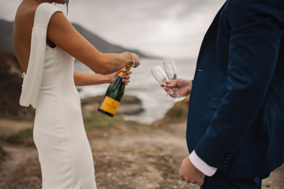 A bride in a white dress opens a champagne bottle while a groom in a blue suit holds two empty glasses, standing outdoors by the seaside with cliffs in the background. by Justin Salem Meyer