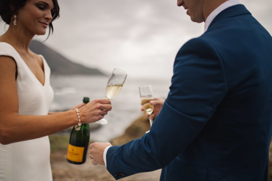 A bride in a white dress pours champagne into a glass held by a groom in a blue suit, standing outdoors by the ocean with a rocky coastline in the background. by Justin Salem Meyer