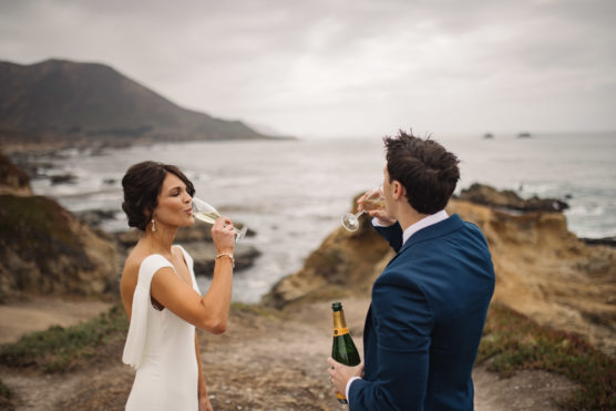 A bride and groom stand on a rocky coastline, facing the ocean, sipping champagne. The bride wears a white dress, and the groom is in a navy suit, holding a champagne bottle. The sky is overcast and mountains are visible in the background. by Justin Salem Meyer