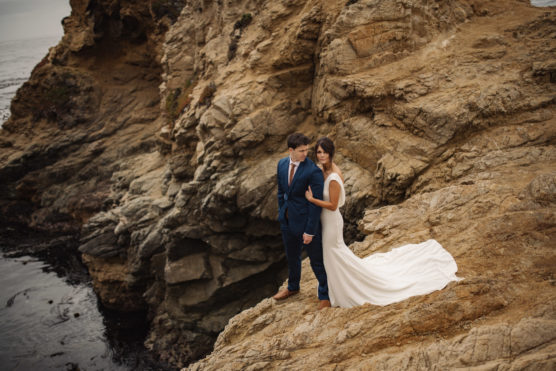 A bride in a white dress and a groom in a blue suit stand close together on a rocky cliff overlooking the ocean, surrounded by textured stone and dramatic coastal scenery. by Justin Salem Meyer