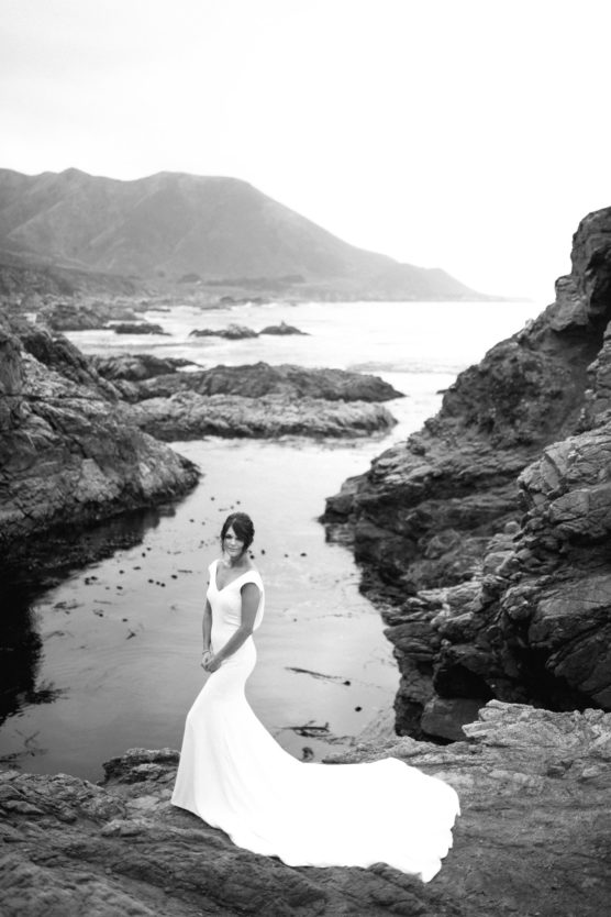 A woman in a long, elegant white dress stands on rocky terrain near a calm body of water, with mountains and coastline in the background under a bright, overcast sky. by Justin Salem Meyer