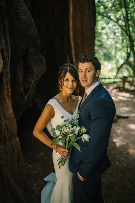 A bride in a white dress holding a bouquet stands beside a groom in a blue suit in a wooded area with sunlight filtering through the trees. Both are smiling and looking at the camera. by Justin Salem Meyer