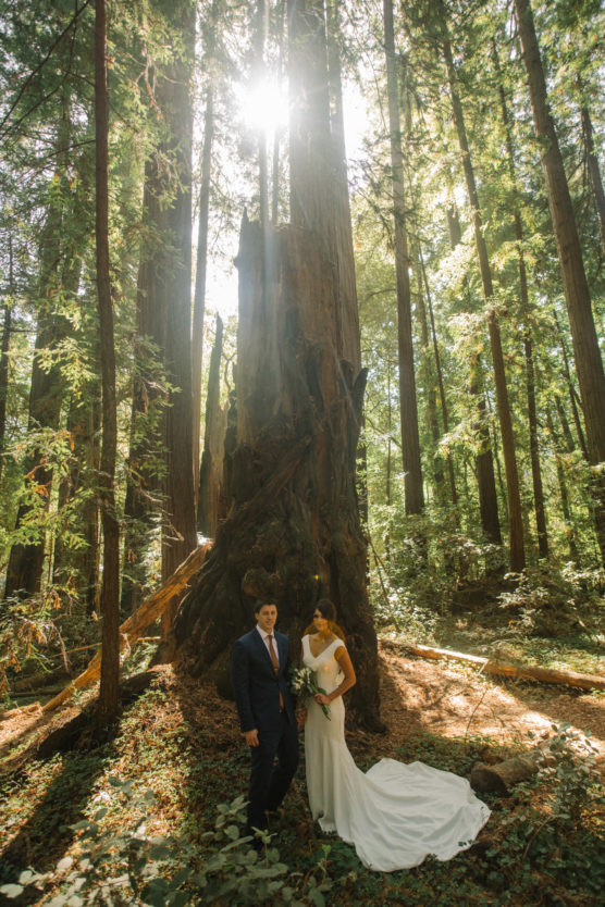 A bride and groom stand together in a sunlit forest, surrounded by tall trees and greenery, with sunlight streaming through the branches behind them. The bride wears a white dress and holds a bouquet. by Justin Salem Meyer