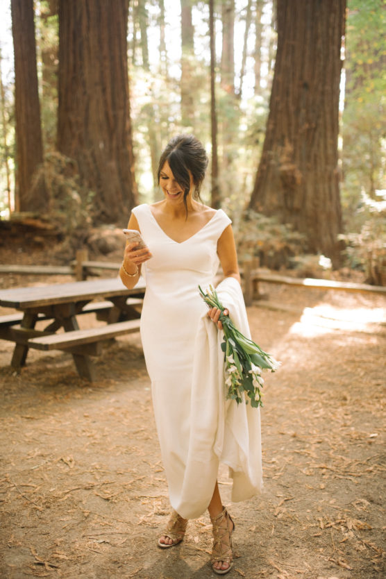 A woman in a white dress, holding a bouquet of flowers, stands in a sunlit forest clearing. She smiles while looking at her phone, with picnic tables and tall trees in the background. by Justin Salem Meyer