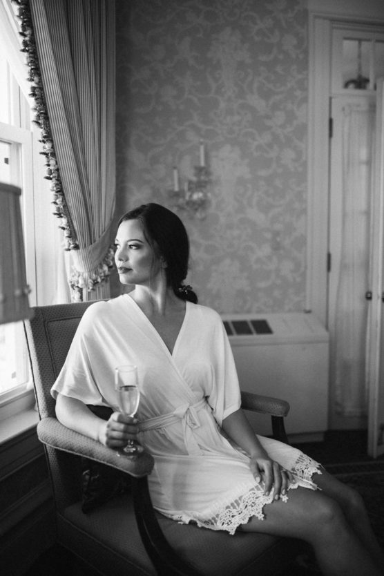 A woman in a white robe sits in an armchair by a window, holding a glass of champagne and gazing outside. The scene is in black and white, with elegant decor and soft natural light. by Justin Salem Meyer
