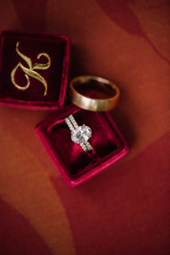 A gold wedding band and a diamond engagement ring are displayed in red velvet ring boxes, one embroidered with a gold letter K, on a red textured surface. by Justin Salem Meyer