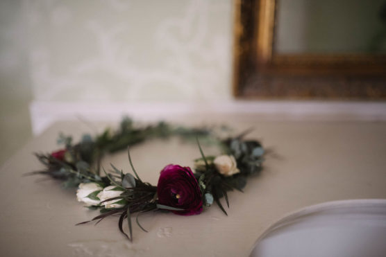 A floral crown with dark pink, white flowers, and green foliage rests on a light-colored surface near a gold-framed mirror. by Justin Salem Meyer