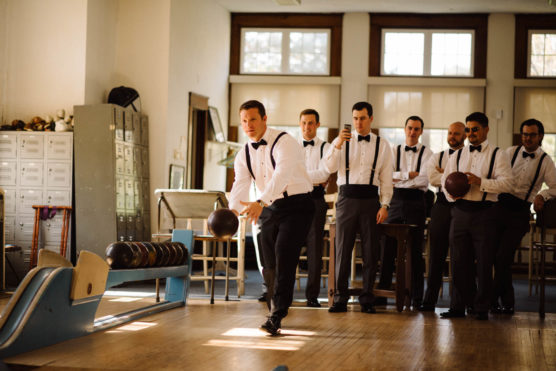 A group of men dressed in formal attire, including suspenders and bow ties, watch as one of them bowls in a vintage-style bowling alley with natural light streaming through the windows. by Justin Salem Meyer