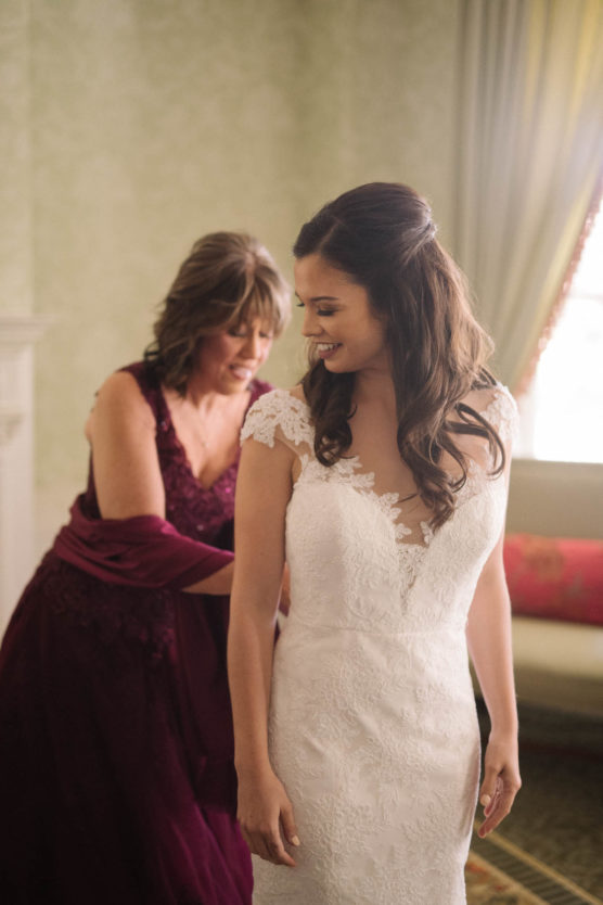 A bride in a white lace wedding dress smiles as a woman in a burgundy gown helps her get ready in a softly lit room with elegant curtains and a vintage-style sofa. by Justin Salem Meyer