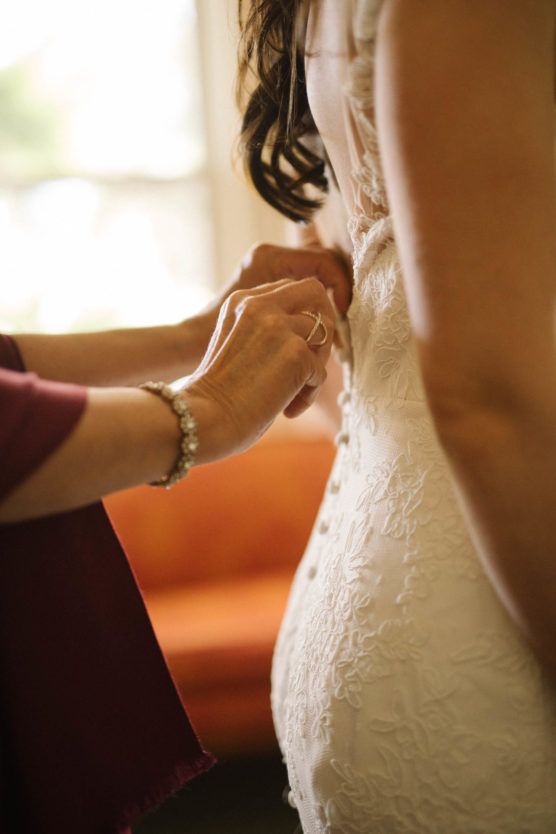 Close-up of someone fastening the buttons on the back of a bride’s lace wedding dress, with soft natural light and an orange sofa blurred in the background. by Justin Salem Meyer