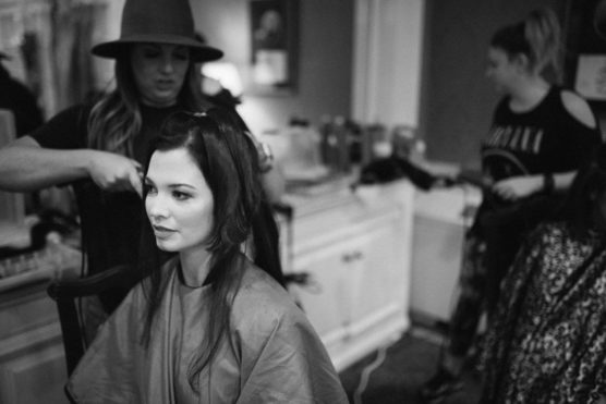 A woman sits in a salon chair with a cape around her shoulders while a hairstylist wearing a hat works on her hair. Another stylist and client are visible in the background. The image is black and white. by Justin Salem Meyer
