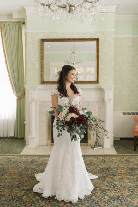 A bride in a white lace gown holds a large bouquet of red, white, and pink flowers with greenery. She stands in a bright, elegant room with a fireplace, chandelier, and green curtains, smiling slightly to the side. by Justin Salem Meyer