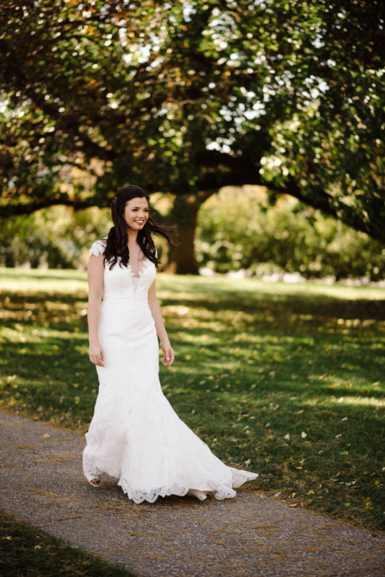 A woman in a white lace wedding dress walks along a path in a sunlit park, surrounded by green grass and large trees with dappled light filtering through the leaves. by Justin Salem Meyer