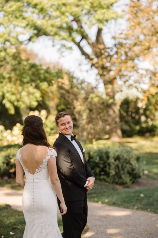 A bride in a white lace dress walks toward a groom in a black suit and bow tie. He is smiling at her as they stand on a garden path surrounded by green trees and bushes. by Justin Salem Meyer
