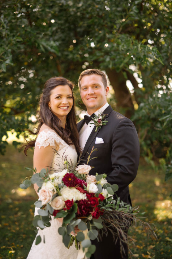 A bride in a white lace dress and a groom in a black tuxedo smile together outdoors, holding a bouquet of red, pink, and white flowers with greenery, with trees and sunlight in the background. by Justin Salem Meyer