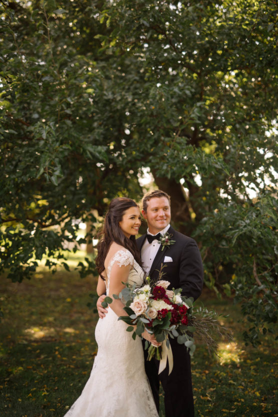 A bride in a white lace dress and a groom in a black tuxedo smile and pose together outdoors, holding a bouquet of white and burgundy flowers, with green trees in the background. by Justin Salem Meyer