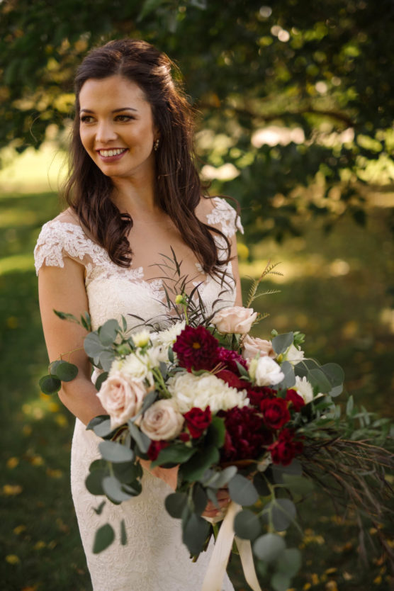 A smiling bride in a white lace wedding dress holds a large bouquet of white, blush, and deep red flowers with greenery, standing outdoors with trees and sunlight in the background. by Justin Salem Meyer