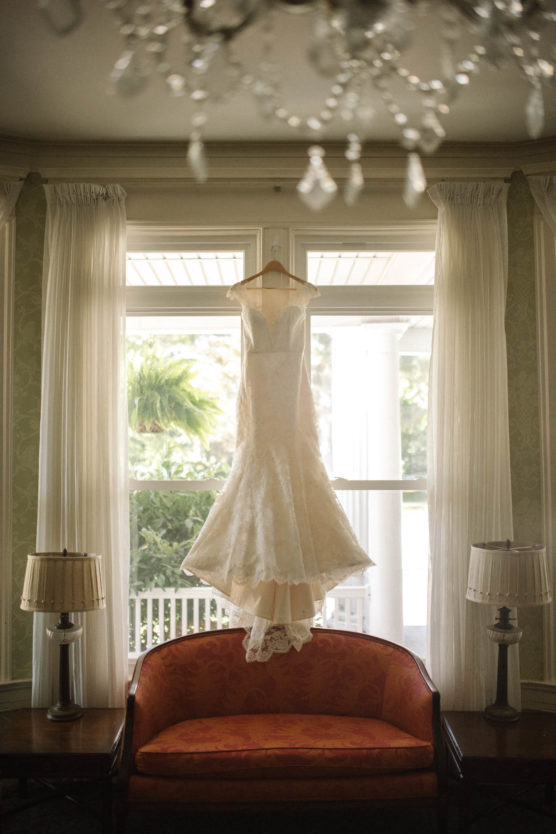 A white lace wedding dress on a hanger is displayed in front of a bright window, above an orange vintage sofa, with two table lamps on either side and a chandelier visible at the top of the image. by Justin Salem Meyer