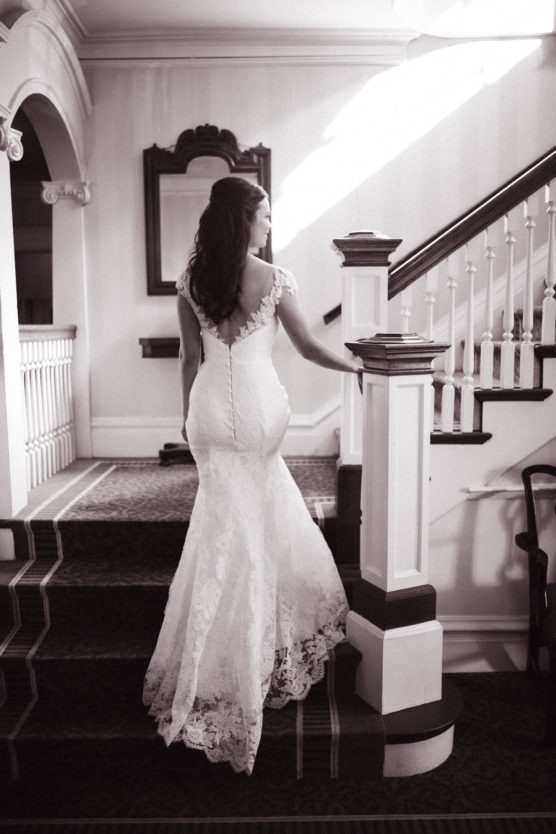 A woman in a lace wedding dress stands on a carpeted staircase, facing away, with light streaming through a window, creating a soft, elegant, and classic atmosphere. by Justin Salem Meyer
