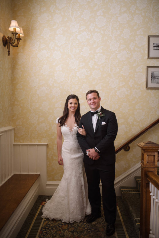 A bride in a white lace dress and a groom in a black tuxedo stand together, smiling, on a staircase with yellow patterned wallpaper and framed pictures on the wall. by Justin Salem Meyer