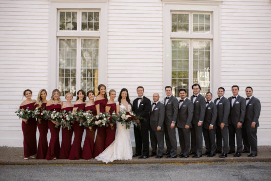 A wedding party poses outdoors in front of a white building. The bride stands in the center in a white dress, flanked by bridesmaids in burgundy gowns and groomsmen in gray suits with bow ties, all smiling. by Justin Salem Meyer