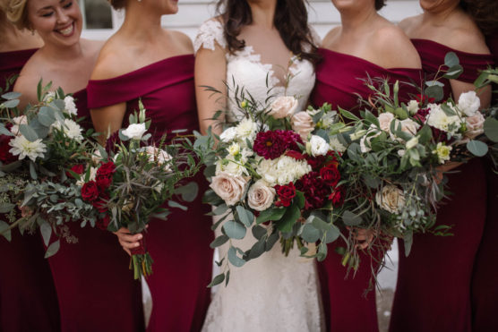 Five women wearing off-the-shoulder burgundy dresses and holding bouquets of red, white, and pink flowers stand around a bride in a white lace gown. Faces are partially visible, focusing on dresses and bouquets. by Justin Salem Meyer