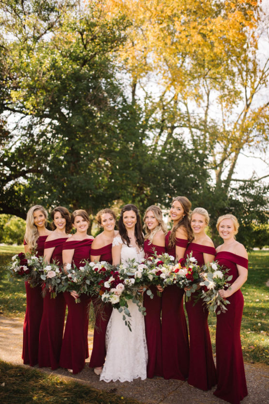A bride in a white dress stands outdoors with eight bridesmaids in matching burgundy dresses, all holding bouquets of flowers. Trees with autumn foliage are in the background, and sunlight filters through the leaves. by Justin Salem Meyer