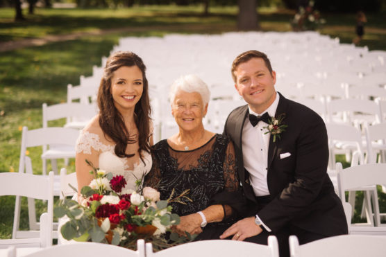 A bride, groom, and elderly woman sit together smiling on white chairs outdoors. The bride holds a bouquet of red and white flowers. The groom wears a tuxedo; the bride wears a white gown. The setting is sunny with greenery in the background. by Justin Salem Meyer