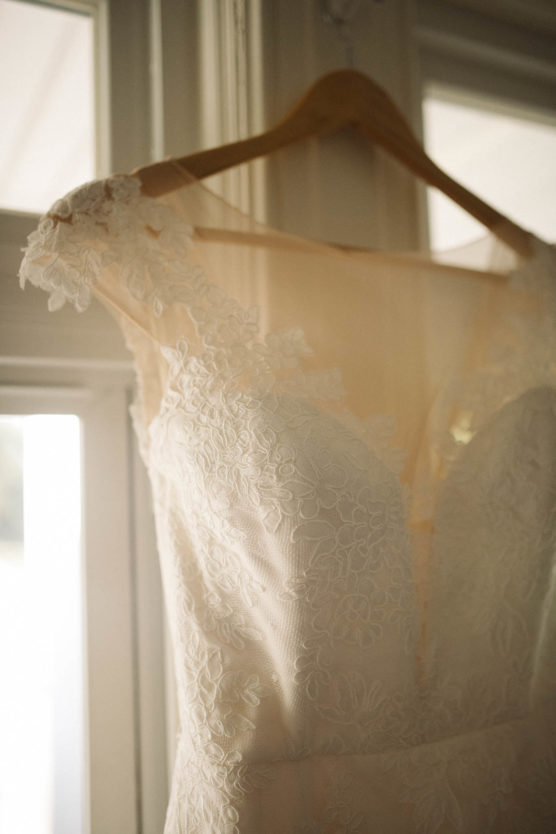A close-up of an elegant lace wedding dress with floral patterns hanging on a wooden hanger near a window, softly lit by natural light. by Justin Salem Meyer