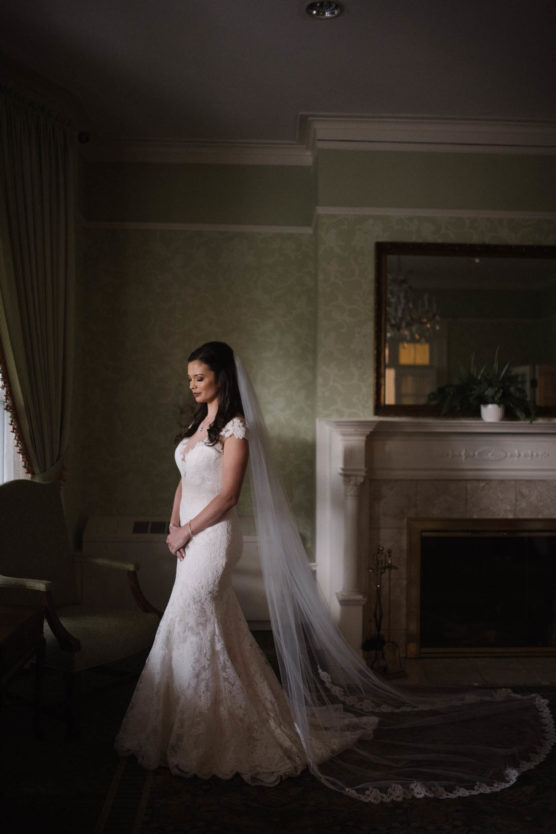 A bride in a white lace gown and long veil stands indoors near a fireplace, hands clasped, looking down softly. The room is softly lit with elegant decor and a large mirror above the mantel. by Justin Salem Meyer