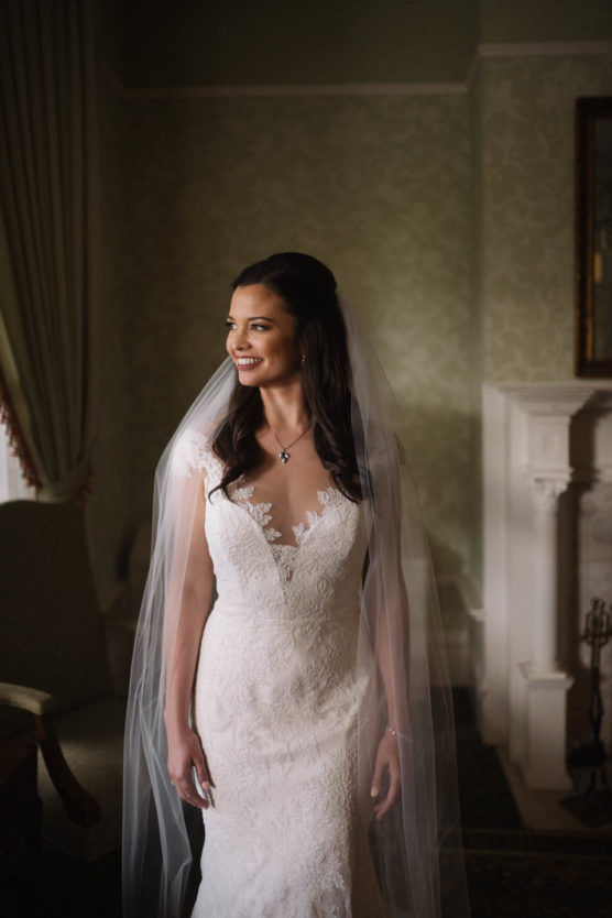 A bride in a lace wedding dress with a long veil stands indoors, smiling and looking to her left. She has long, dark hair and wears a necklace, with soft light illuminating her. Elegant decor is visible in the background. by Justin Salem Meyer