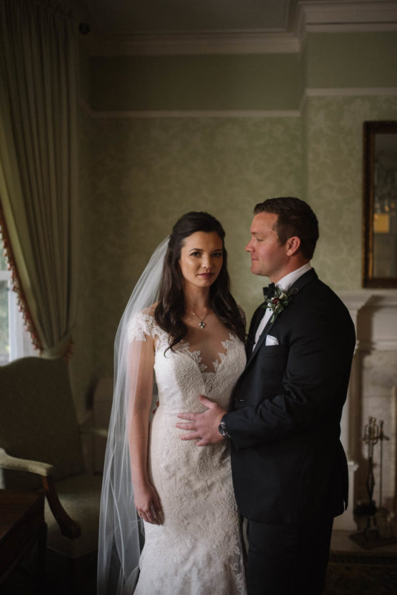 A bride in a lace wedding dress and veil stands beside a groom in a black tuxedo. They pose indoors in an elegant room with green walls, a fireplace, and a curtained window. The groom gently holds the bride’s waist. by Justin Salem Meyer