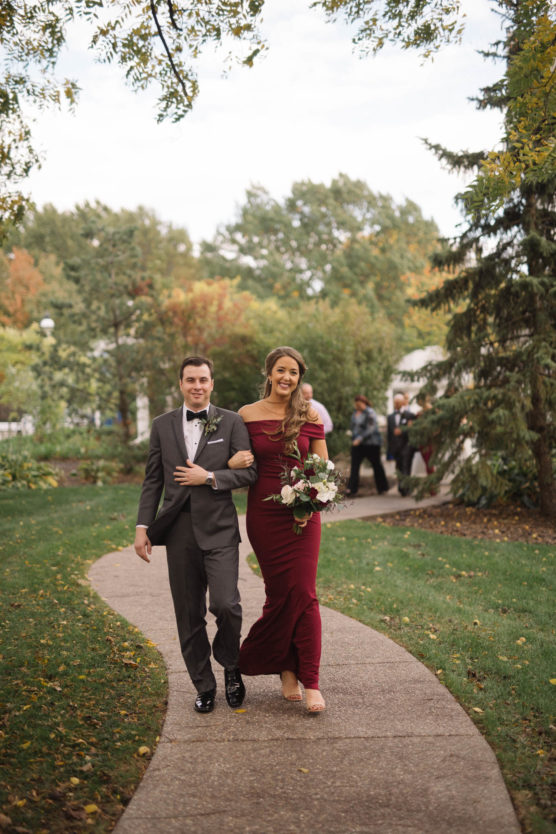 A man in a gray suit and a woman in a burgundy dress walk arm-in-arm along a curved garden path, smiling, surrounded by green trees and grass on a cloudy day. She holds a bouquet of flowers. by Justin Salem Meyer