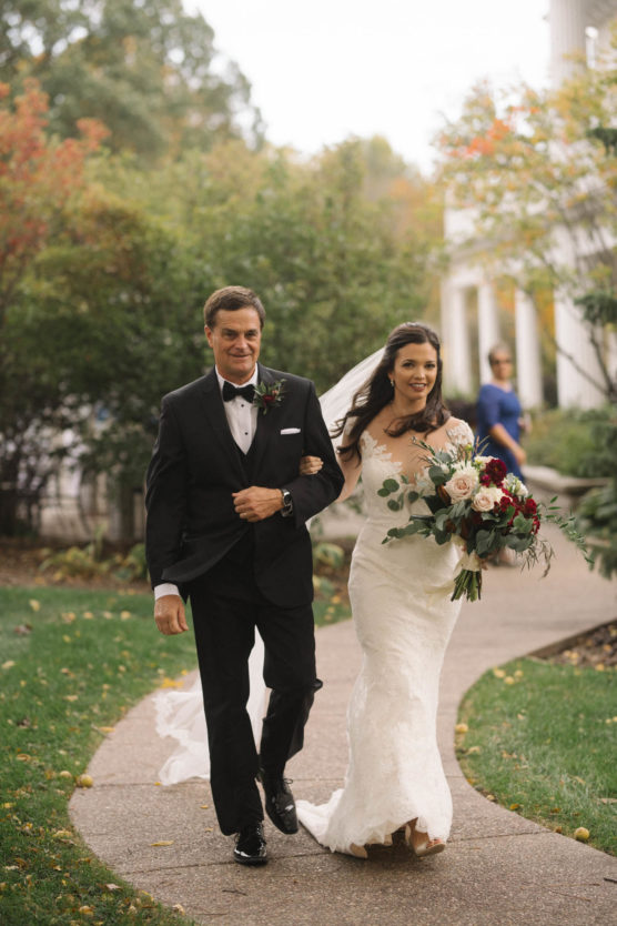 A bride in a white dress holding a bouquet walks arm-in-arm with a man in a black tuxedo down a garden path, both smiling. Green trees and autumn foliage are visible in the background. by Justin Salem Meyer