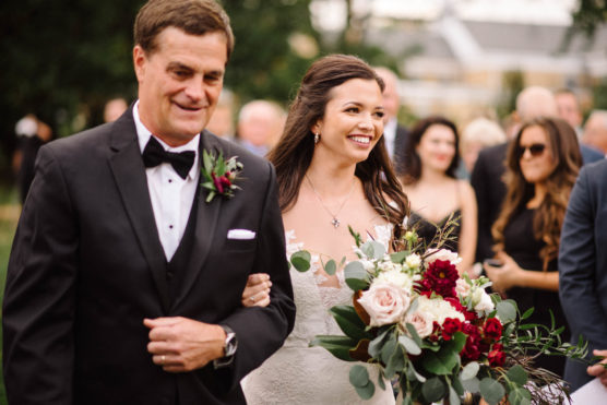 A smiling bride in a white dress holding a bouquet walks arm in arm with a man in a tuxedo at an outdoor wedding, with guests and greenery in the background. by Justin Salem Meyer