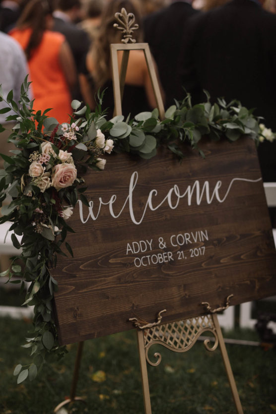 A wooden sign decorated with greenery and pink flowers reads, Welcome. Addy & Corvin. October 21, 2017. The sign is displayed on an easel at an outdoor event with people gathered in the background. by Justin Salem Meyer