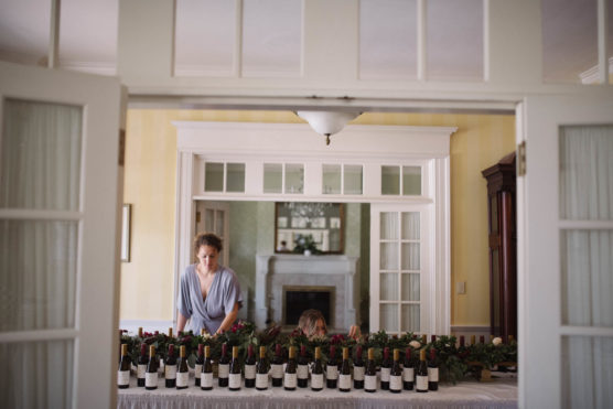 A person stands behind a long table covered with wine bottles, with another person partially visible beside them. The room is bright with white trim, yellow walls, and a fireplace in the background. by Justin Salem Meyer
