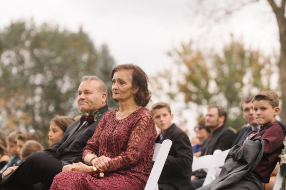 A woman in a maroon lace dress sits attentively beside a man in a dark suit at an outdoor event, surrounded by others seated on white chairs. Trees with autumn leaves are in the background. by Justin Salem Meyer