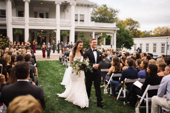 A bride and groom, both smiling, walk down the aisle outdoors after their wedding ceremony. Guests seated on white chairs watch as the couple passes by, with a large white building in the background. by Justin Salem Meyer