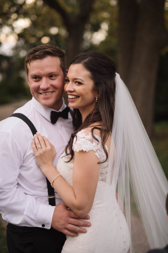 A bride in a white lace gown and veil smiles beside a groom in a white shirt with suspenders and a black bow tie; they stand outdoors, looking happy on their wedding day. by Justin Salem Meyer