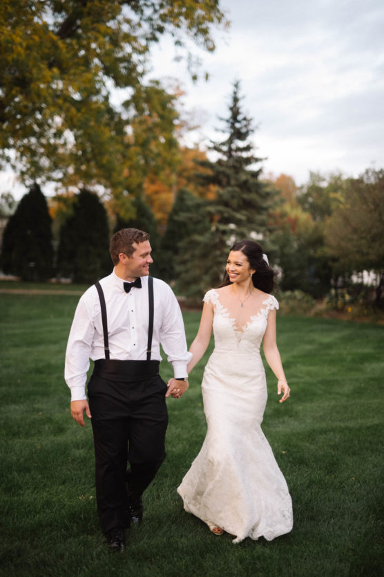 A bride in a white lace gown and a groom in black suspenders and a bow tie walk hand in hand on green grass, smiling at each other, with trees and a cloudy sky in the background. by Justin Salem Meyer