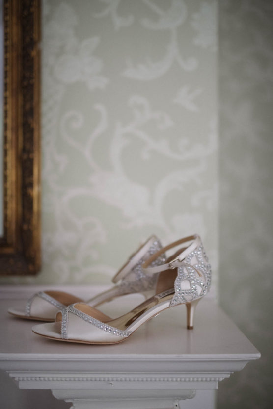 A pair of elegant white open-toe heeled shoes with rhinestone embellishments sits on a white ornate table against a pale floral-patterned wallpaper. by Justin Salem Meyer