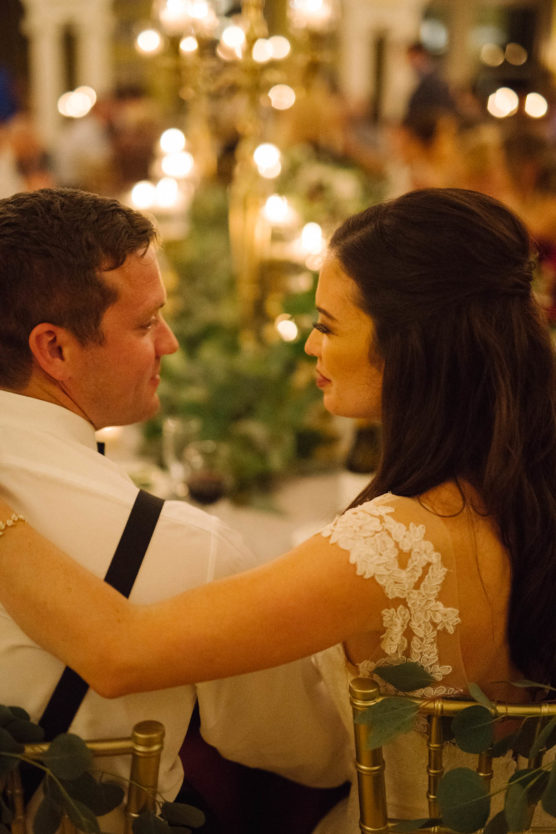 A bride and groom sit close together at a candlelit wedding reception table, looking into each others eyes and smiling warmly, surrounded by greenery and softly glowing lights. by Justin Salem Meyer