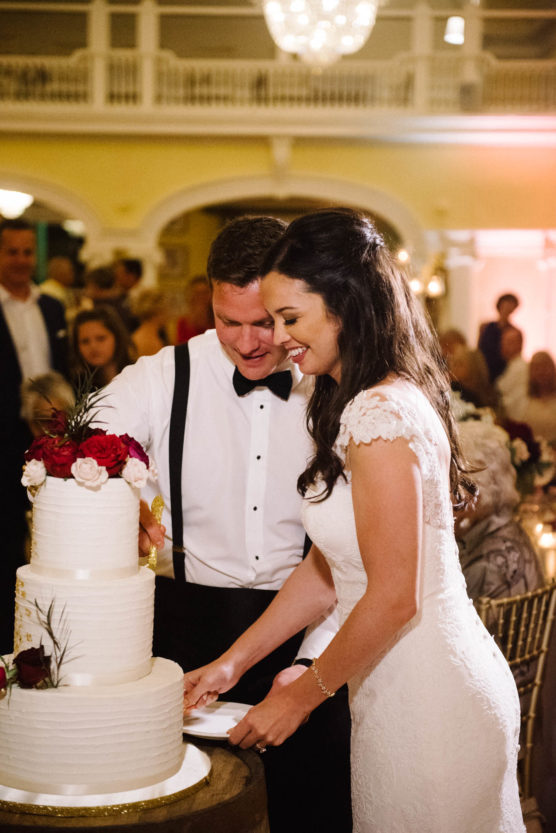 A bride and groom, dressed in formal wedding attire, smile as they cut a white, tiered wedding cake decorated with red and white flowers, surrounded by guests in an elegant, well-lit reception hall. by Justin Salem Meyer