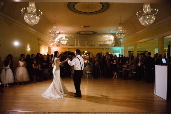 A bride and groom share their first dance in an elegant ballroom, surrounded by guests watching and standing near tables. Chandeliers hang from the ceiling, illuminating the scene with warm light. by Justin Salem Meyer