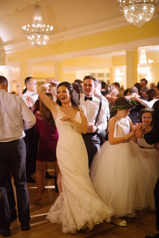 A bride in a white dress dances joyfully among guests at a wedding reception, surrounded by people in formal attire under sparkling chandeliers in a warmly lit room. by Justin Salem Meyer