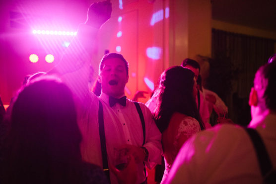 A group of people dance at a party, bathed in pink and purple lights. A man in a white shirt with suspenders and a bow tie raises his arm and sings or shouts joyfully, surrounded by others. by Justin Salem Meyer