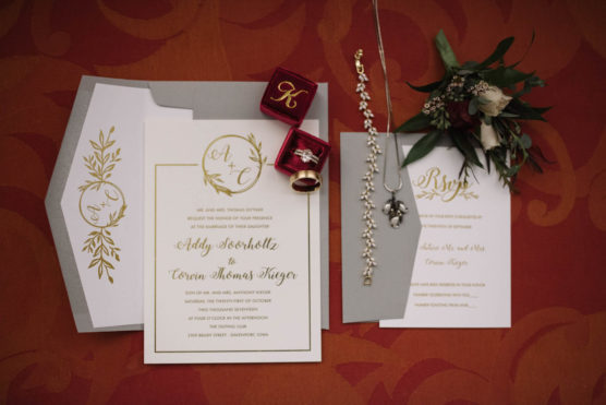 Elegant wedding invitation suite with gold and white cards, a red ring box holding rings, a necklace, a bracelet, and a small flower boutonniere arranged on a red patterned surface. by Justin Salem Meyer
