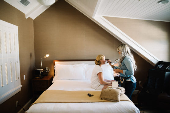 A woman sits on a bed in a cozy attic bedroom while another woman applies her makeup. The room has slanted ceilings, a lamp on a bedside table, and folded towels and a makeup bag on the bed. by Justin Salem Meyer